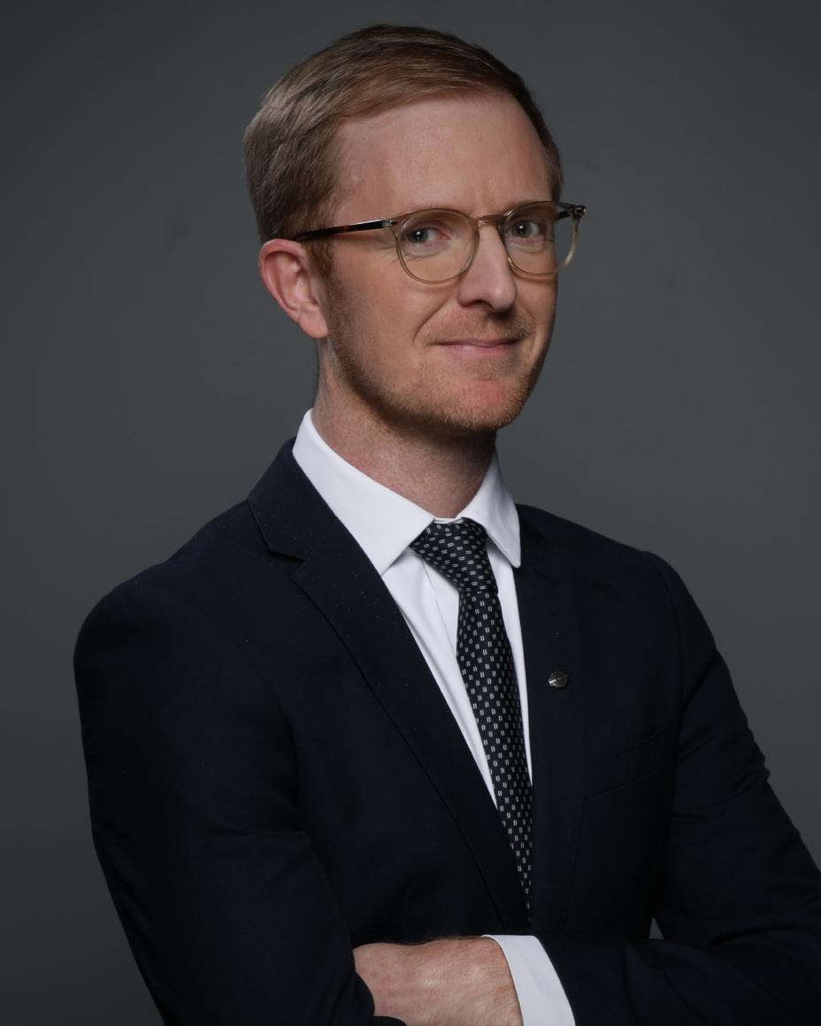 Dominic smiling in a black suit against a gray background.
