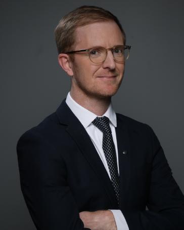 Dominic smiling in a black suit against a gray background.