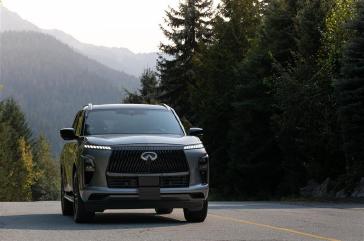 A grey 2025 INFINITI QX80 driving on a forested mountain road with yellow dividing
lines, surrounded by tall evergreen trees and misty mountains in the background