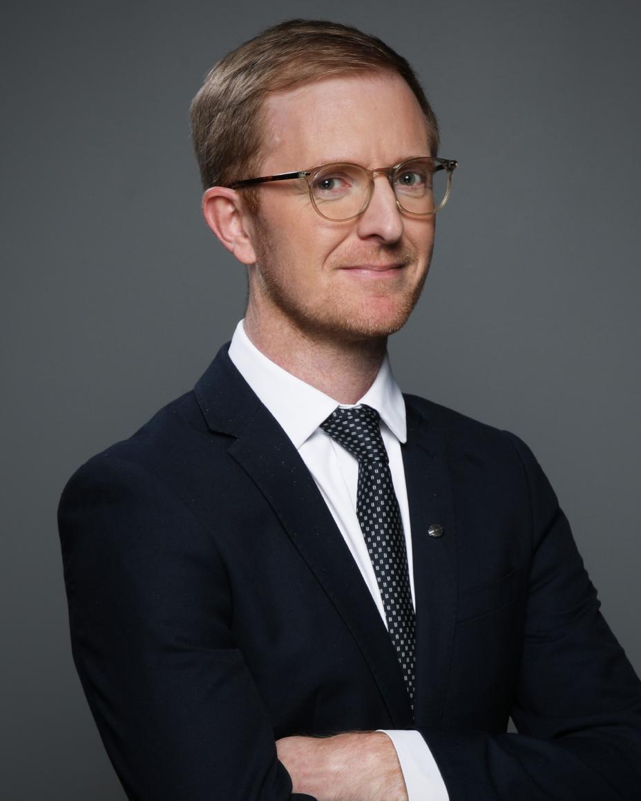 Dominic smiling in a black suit against a gray background.