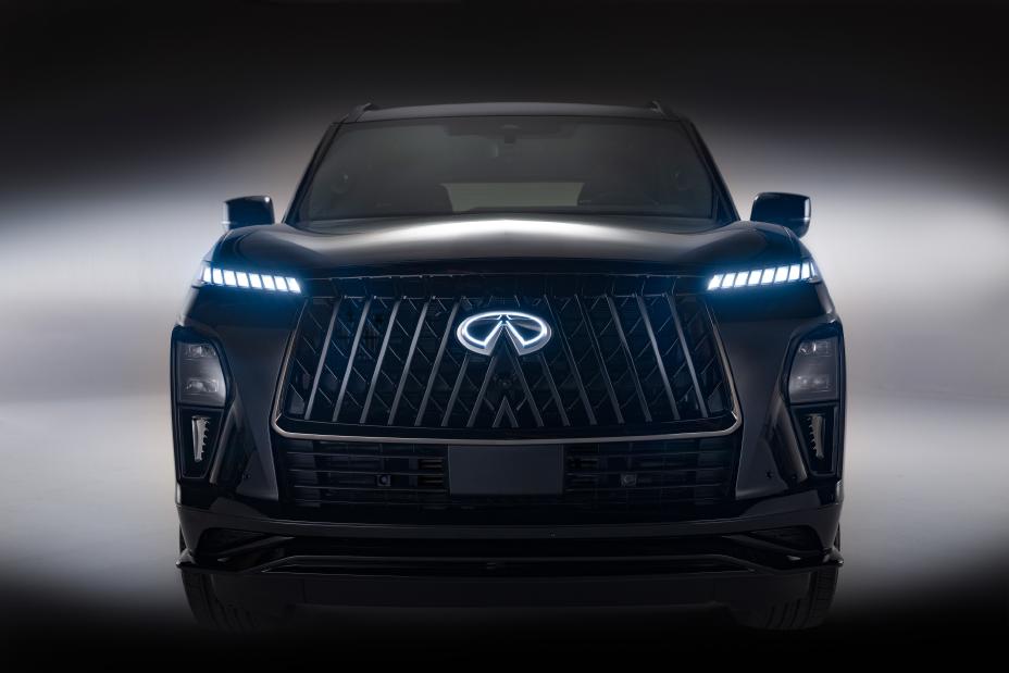 Head-on view of a black Infiniti SUV with LED headlights turned on, showcasing a bold grille with the Infiniti emblem; the dark background enhances the vehicle’s illuminated front design