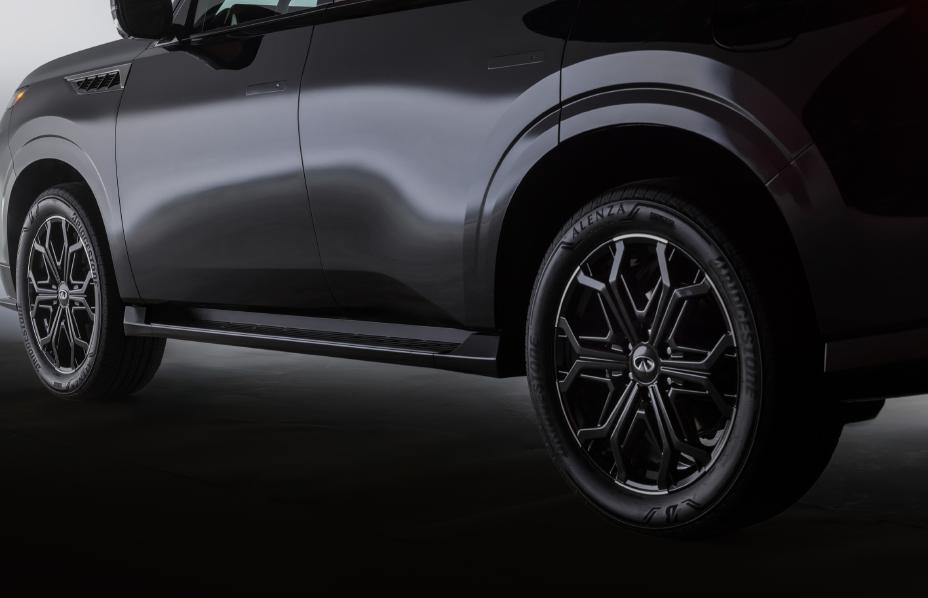 Close-up of the side profile of the 2026 INFINITI QX80, showing the front and rear wheels with Venezia-branded tires and intricate alloy wheel designs. The dark-coloured body features sleek lines and reflective surfaces.