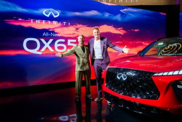 Two men pose next to the 2027 INFINITI QX65, a QX65 stage display is behind them.
