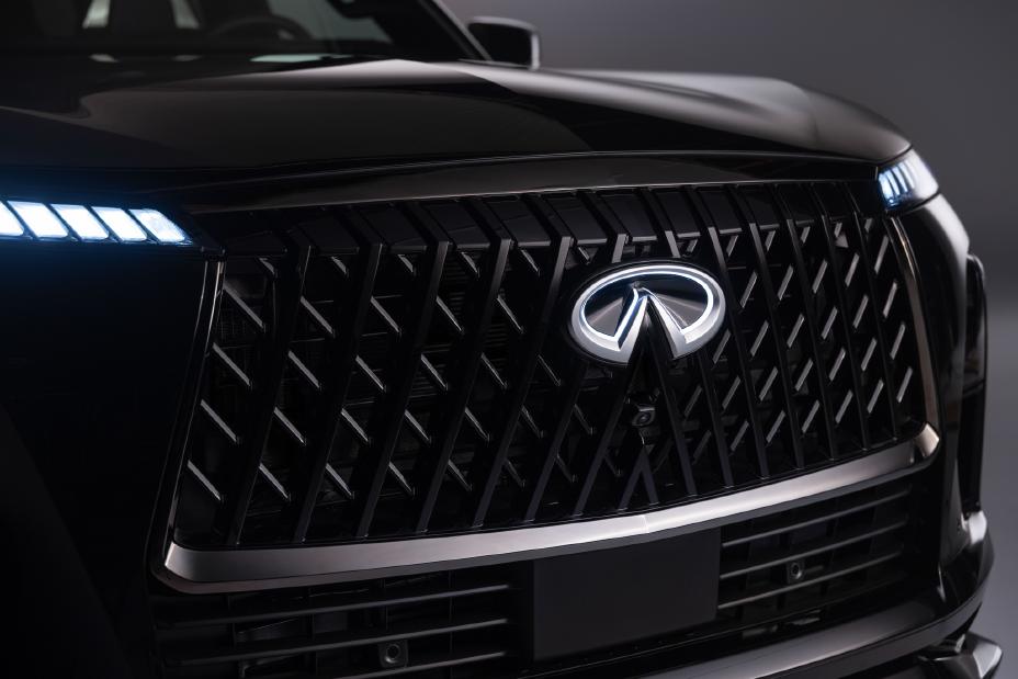 Front grille of an Infiniti vehicle with a diamond-patterned mesh design and a prominent Infiniti logo in the center; LED headlights are illuminated on both sides.