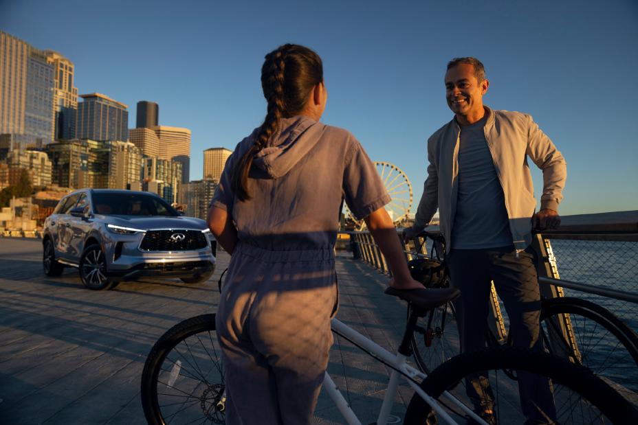 Padre e hija con bicicletas, de espaldas al lago en un entorno urbano; el papá sonríe a su hija mientras la INFINITI QX60 está estacionada al fondo.