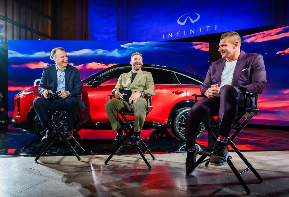 Three men sit in chairs, talking, in front of the 2027 INFINITI QX65 on a stage.