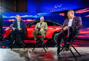 Three men sit in chairs, talking, in front of the 2027 INFINITI QX65 on a stage.
