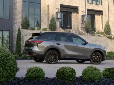 Angled right profile view of the 2026 INFINITI QX60 SPORT parked in front of a brick house with lush landscaping.