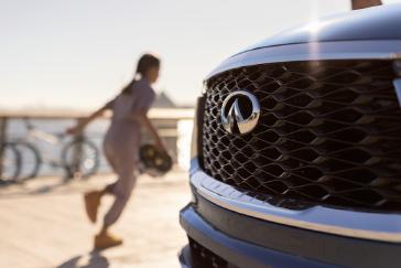 Primer plano de la parrilla frontal de un vehículo INFINITI con su emblema, mientras una persona pasa caminando al fondo cerca del mar con luz de atardecer.