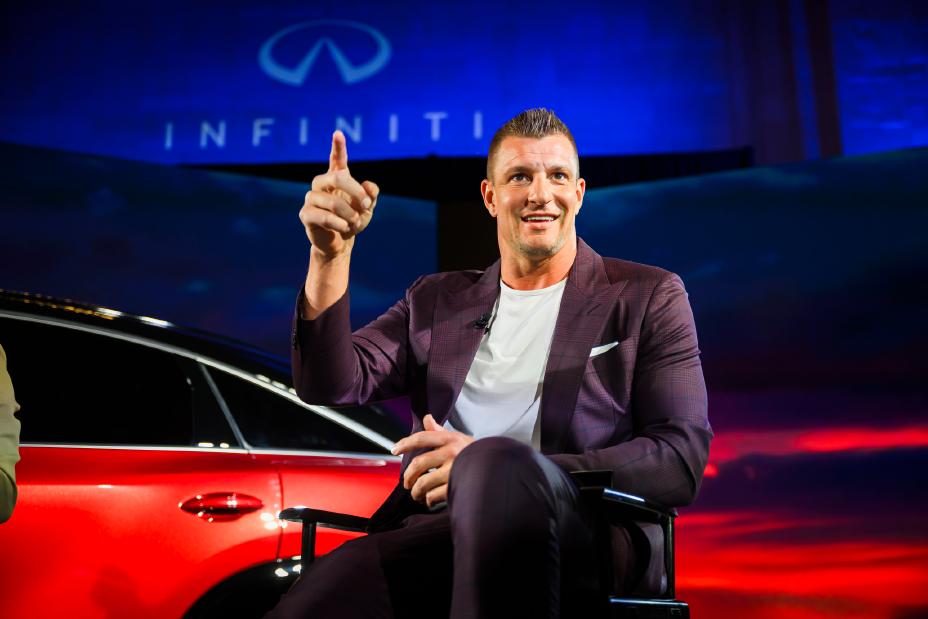 Rob Gronkowski sits in front of the 2027 INFINITI QX65, he is pointing out towards a crowd.