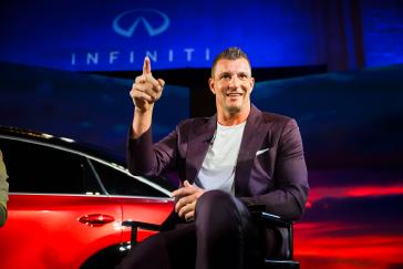 Rob Gronkowski sits in front of the 2027 INFINITI QX65, he is pointing out towards a crowd.