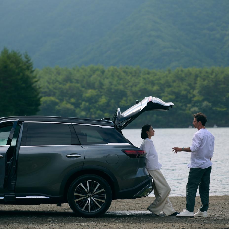 Pareja junto a una INFINITI QX60 estacionada frente a un lago en medio de un bosque; ella recostada en la parte posterior de la SUV y él de frente, disfrutando un momento de descanso y conexión con la naturaleza.
