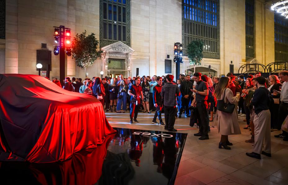 The 2027 INFINITI QX65 hidden under a large red cloth, people bustle about around the display.
